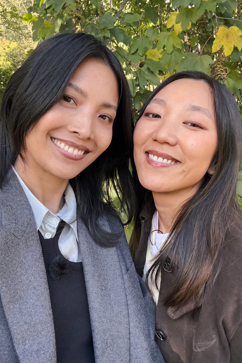 Due donne sorridenti, una con un cappotto grigio sopra un maglione nero, l'altra con un cappotto marrone scuro sopra una camicia bianca; sfondo di fogliame verde.