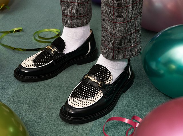 Sorte og hvite loafers med perforert design, med en metallkjede aksent foran. Brukt med grå rutete bukser og hvite sokker.