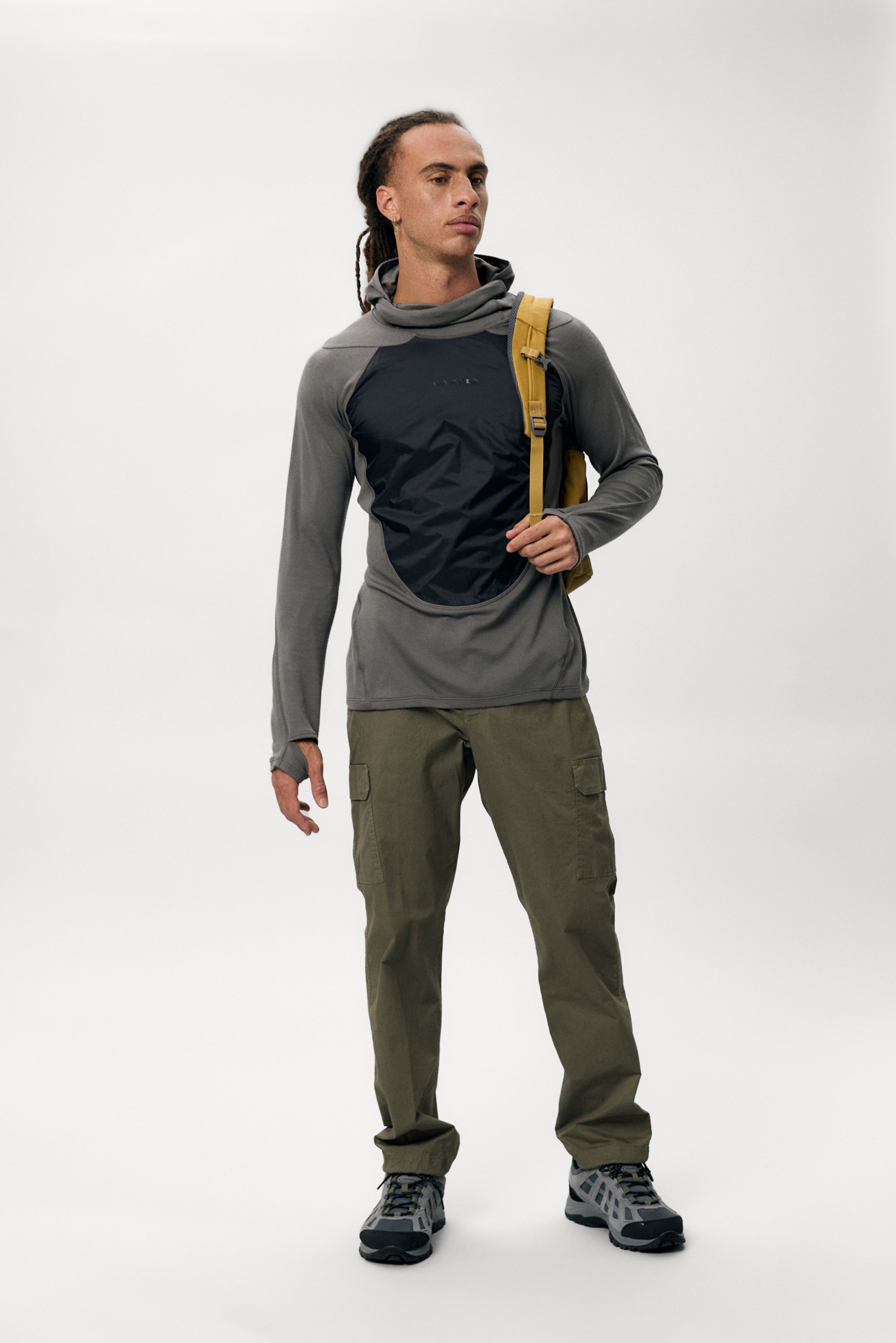 A man with dreadlocks wears a gray and black top, green cargo pants, and a yellow backpack, posed against a plain white background.