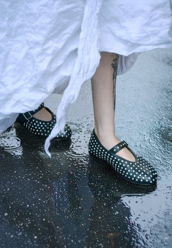 Schwarze Mary-Jane-Schuhe mit silbernen Nieten, runder Spitze und einem Knöchelriemen, auf einem nassen Gehweg, teilweise von einem weißen Kleidungsstück verdeckt.
