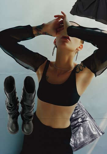 Une personne avec les cheveux courts pose devant un fond neutre, portant un top court noir et des manches transparentes, avec des bottes noires et une jupe brillante à proximité.