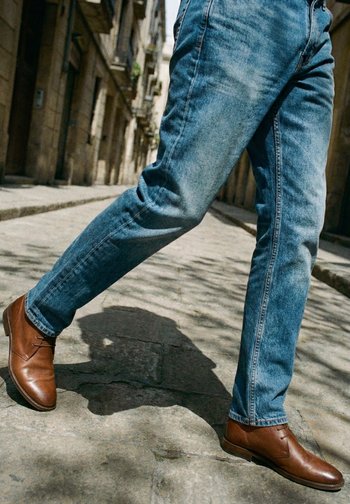 Mann som går på steinbelagt fortau iført brune skinnsko og blå jeans i en solfylt, smal gate med steinbygninger.