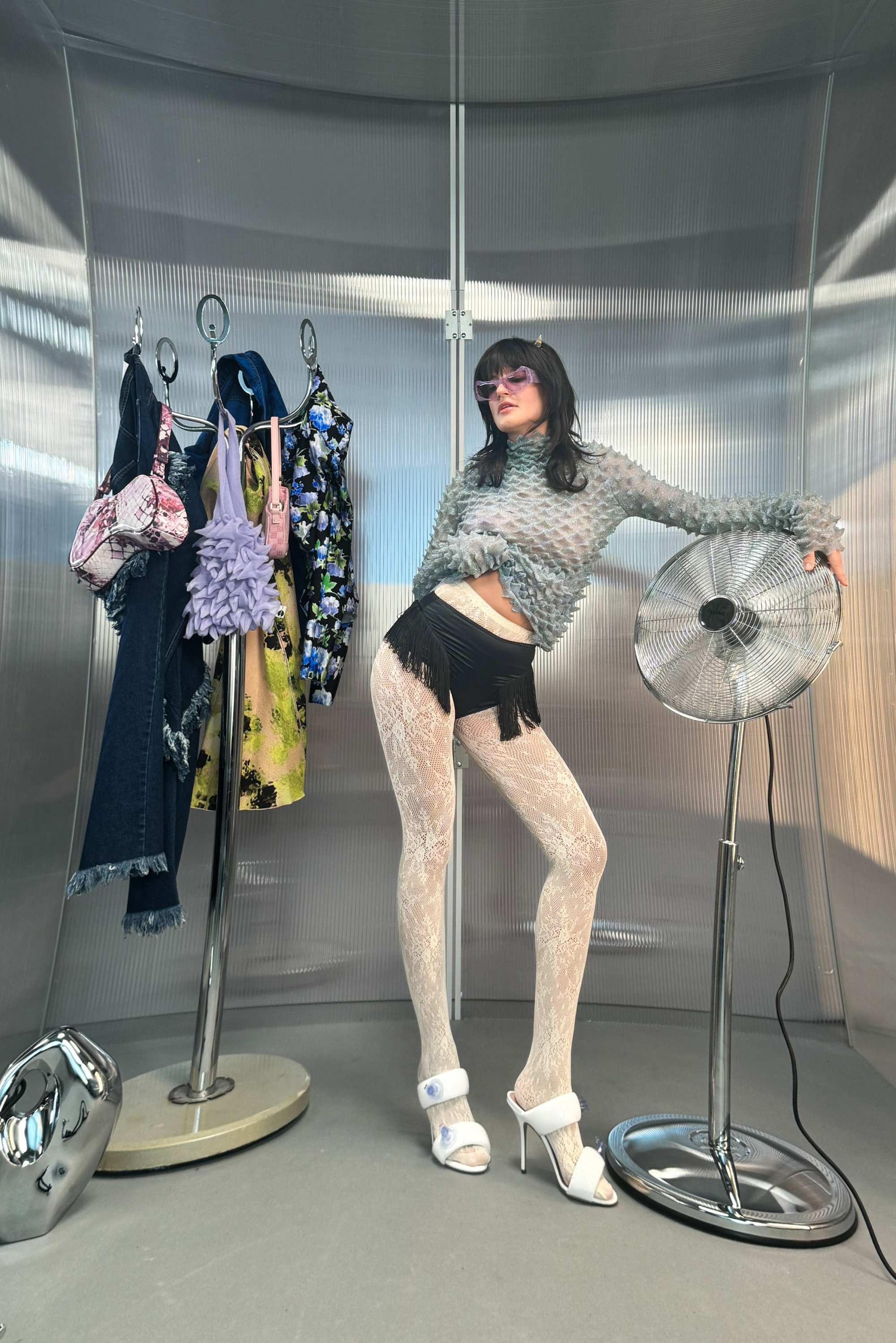 Model poseert in een stijlvolle outfit met franjeshorts en gestreepte panty's, omringd door kleurrijke kleding aan kledinghangers en een ventilator.