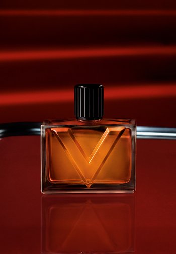 Flacon de parfum rectangulaire ambré avec un bouchon noir strié et un grand V embossé sur le devant, posé sur un fond rouge foncé avec une surface réfléchissante.