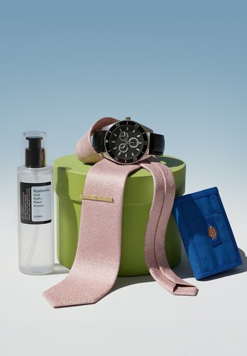 Klare Flasche mit Hyaluronsäure-Serum, schwarze Armbanduhr auf rosa Krawatte, die über eine grüne runde Schachtel gelegt ist, und blauer Geldbeutel auf hellem Hintergrund.