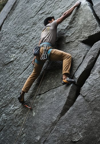 Un climber in pantaloni beige e scarpe da arrampicata nere sta salendo una superficie rocciosa grigia e ruvida, utilizzando prese per le mani e attrezzature da corda per il supporto.