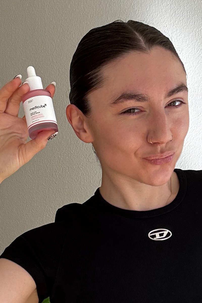 Personne aux cheveux foncés et anneau de nez tient une bouteille rose du produit de soin de la peau Medicube près de son visage, portant un haut noir devant un mur uni.