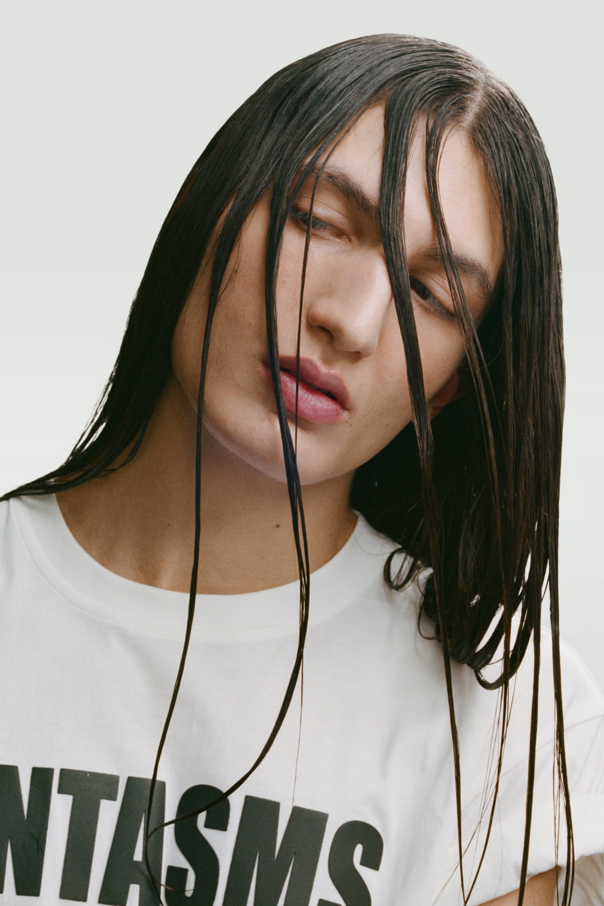 Una persona con el cabello largo y mojado mira hacia abajo, vistiendo una camiseta blanca con texto negro en negrita. El fondo es de color gris claro, lo que realza su expresión.