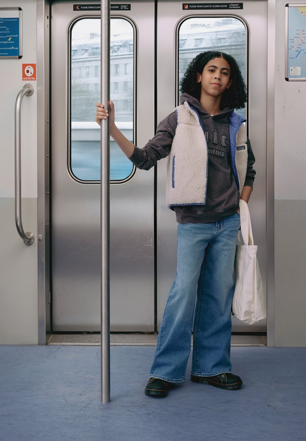 En person står i tunnelbanan och håller i en stång. De har på sig en ljus fleeceväst över en mörk hoodie, med vida blå jeans och svarta skor.