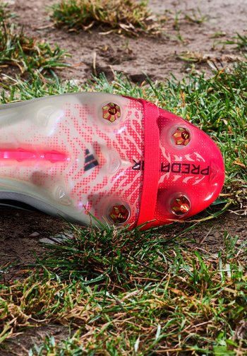Semelle de crampons de football avec des accents rouges et six crampons en métal reposant sur de l'herbe et de la terre, affichant partiellement le nom de la marque.