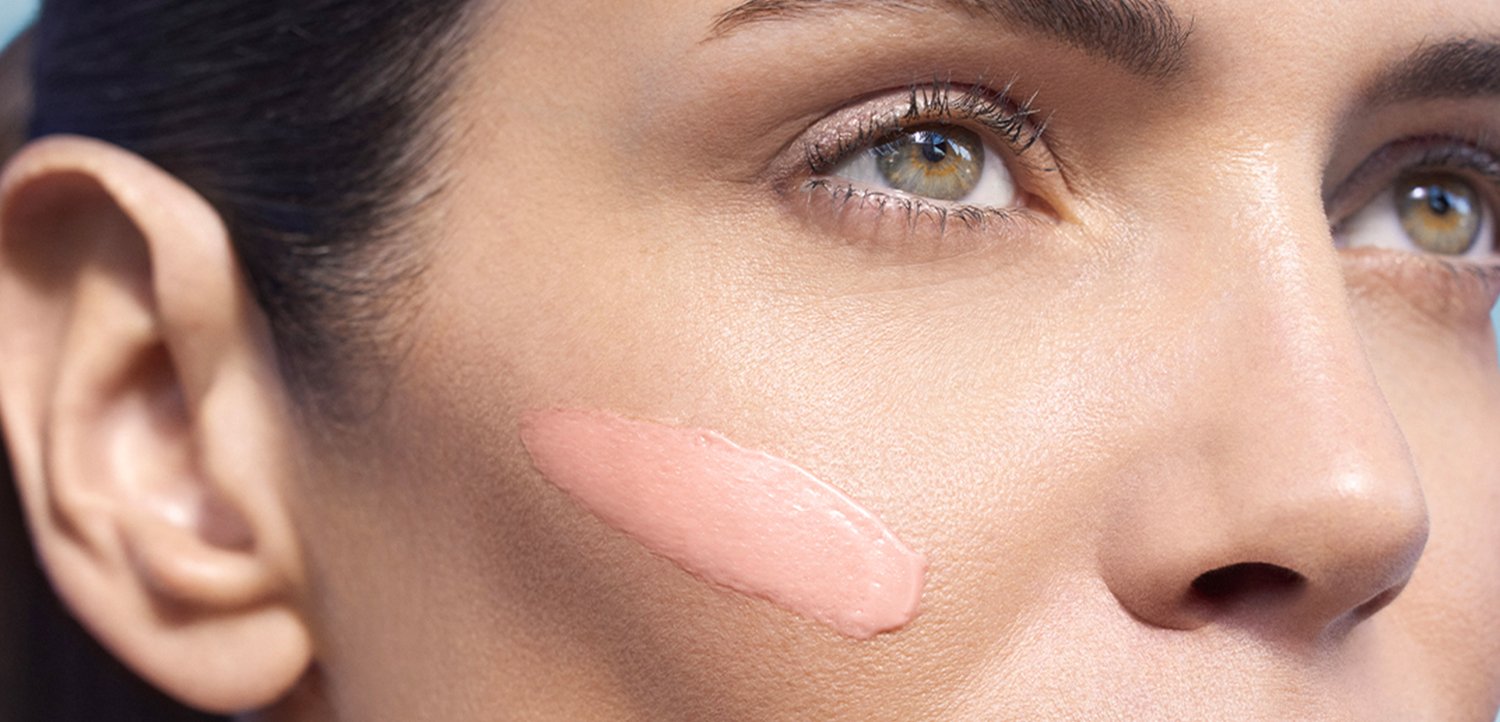 Close-up of a woman's face with a light pink cream swiped across her cheek, emphasizing smooth skin and natural beauty.