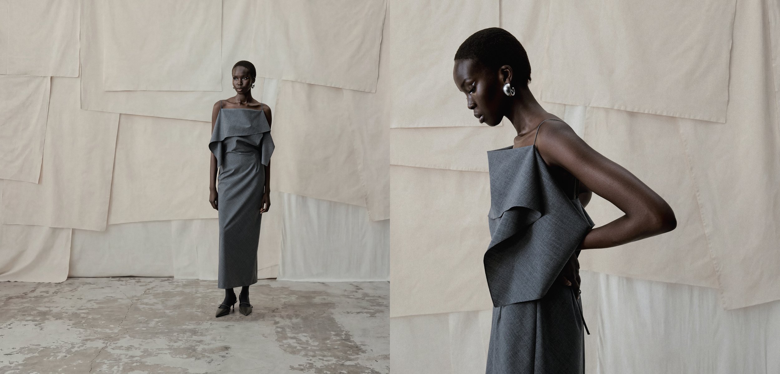 Schwarze Frau, die ein ärmelloses graues Midi-Kleid mit Rüschen am Oberteil und schwarze Schuhe trägt, steht und posiert vor einem beigefarbenen Hintergrund.