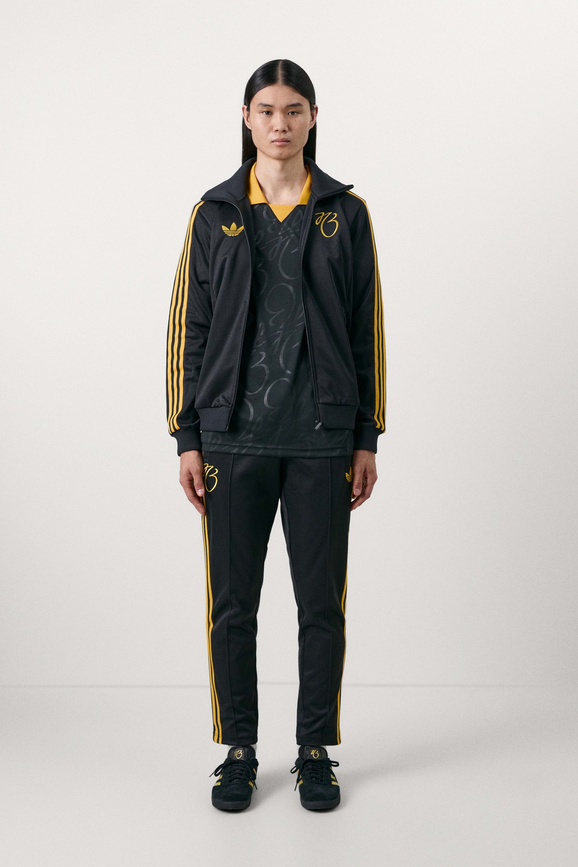 Person wearing a black tracksuit with yellow stripes and logo, standing in a neutral setting, looking directly at the camera.