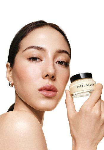 Kobieta z gładką skórą trzyma słoik kremu Bobbi Brown blisko twarzy na białym tle.