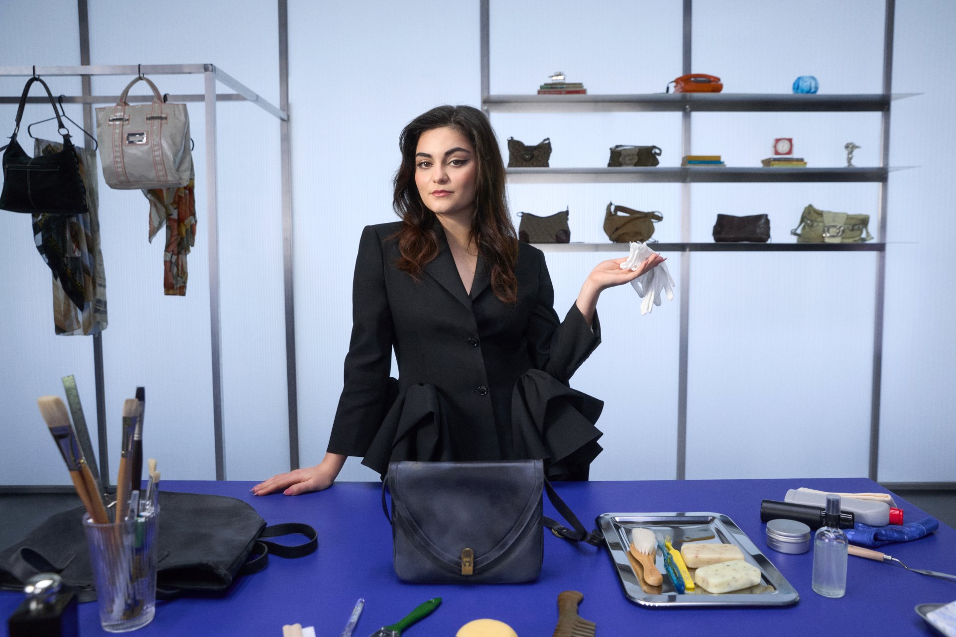 A woman in a stylish black blazer stands beside a table with cleaning tools, bags on shelves behind her, and holds white gloves in one hand.