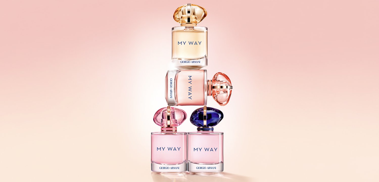 Pila di sei flaconi di profumo Giorgio Armani My Way in diverse tonalità di rosa, viola e oro su uno sfondo morbido pesca.