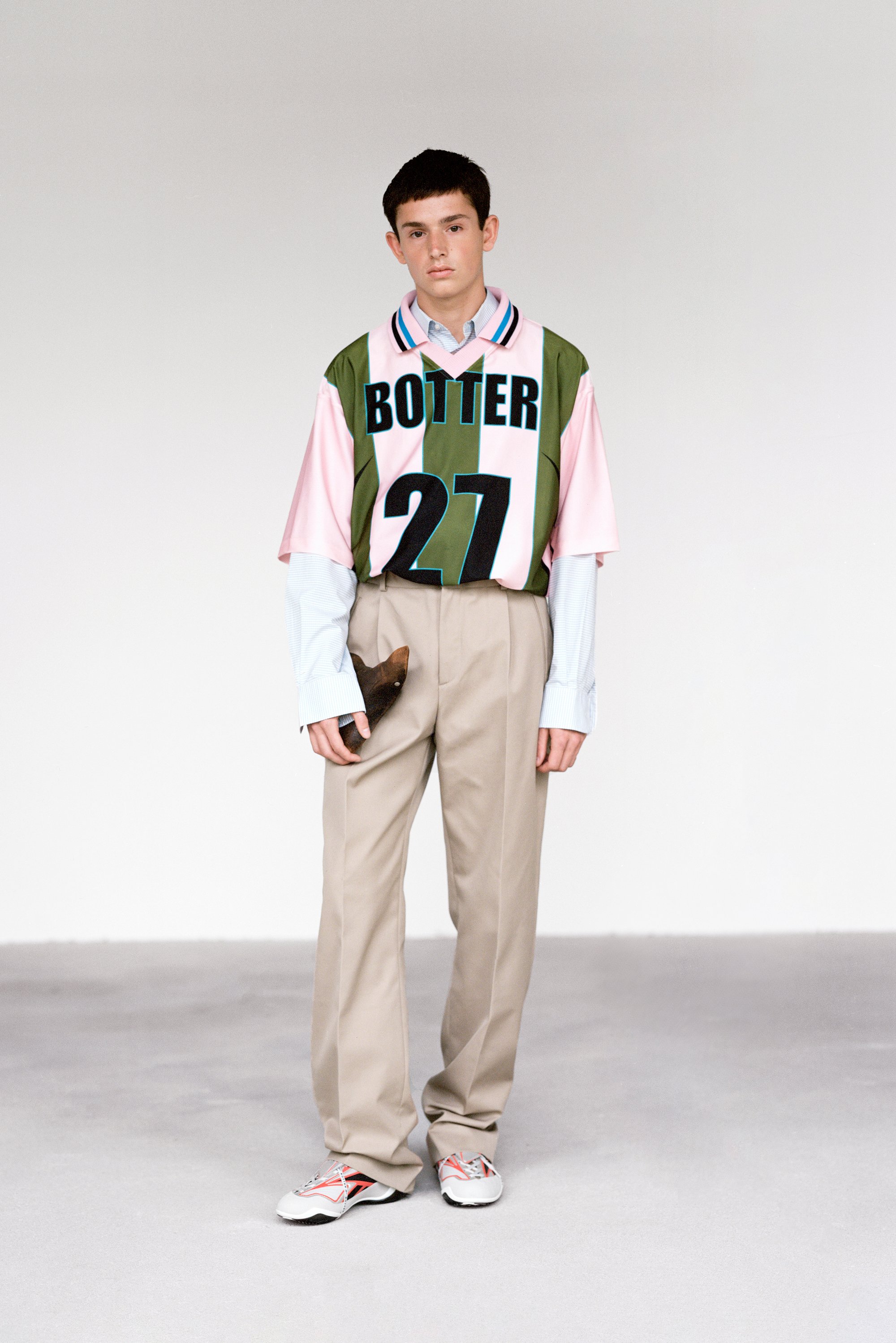Un modelo masculino se encuentra en un espacio minimalista, vistiendo una camiseta verde y rosa con "BOTTER 27", pantalones beige y zapatillas blancas.