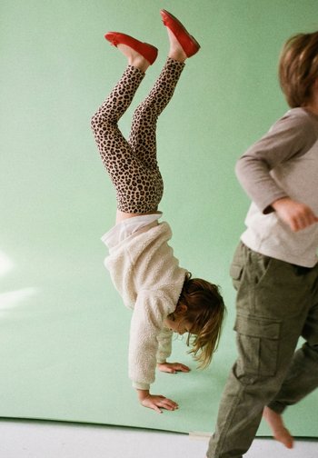 Enfant en legging à imprimé léopard et chaussures rouges faisant un poirier pendant qu'un autre enfant en pantalon cargo bouge à proximité sur un fond vert.