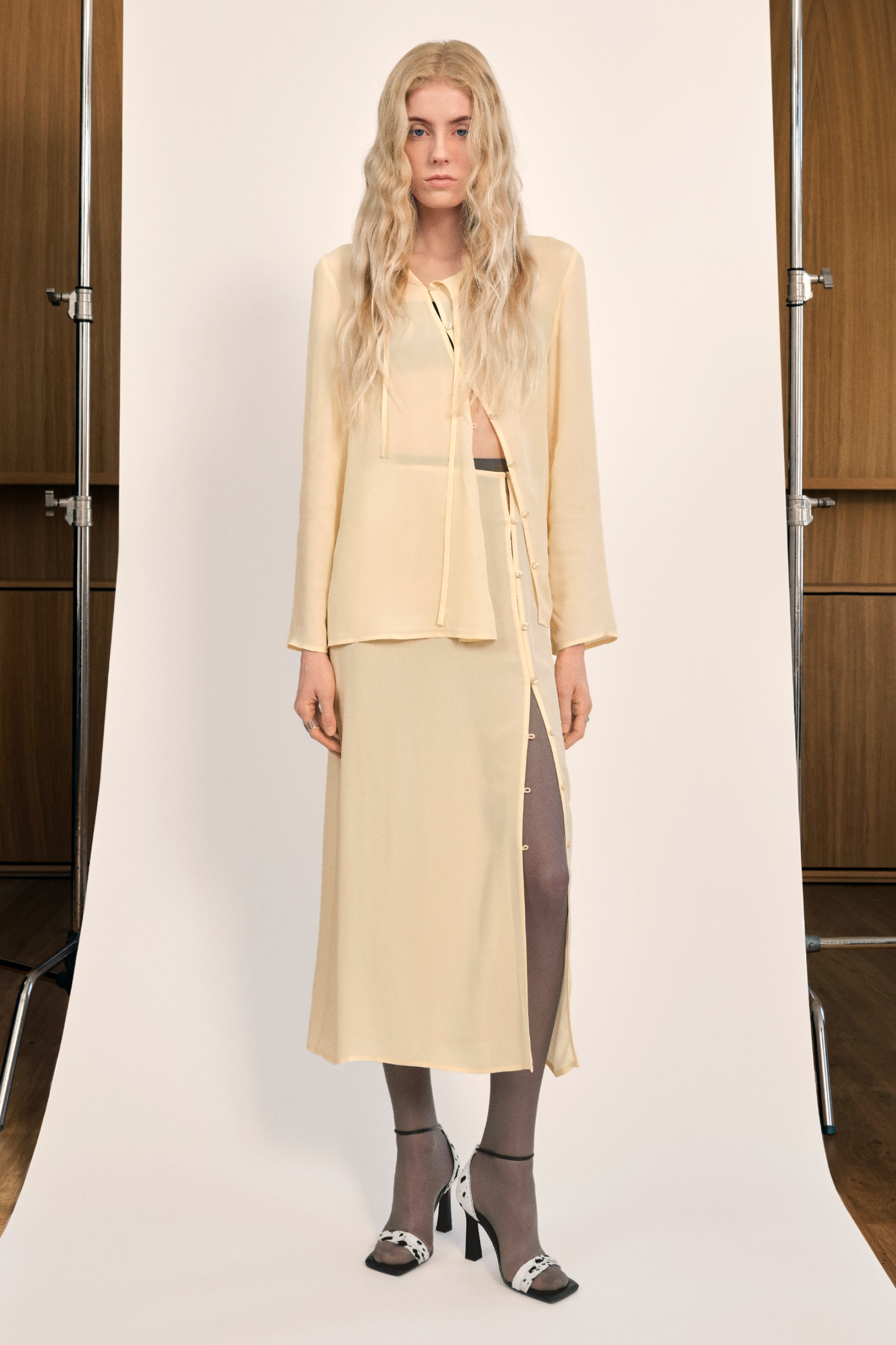 A model stands against a plain backdrop, wearing a beige top and long skirt with buttons, paired with patterned heels and gray tights.