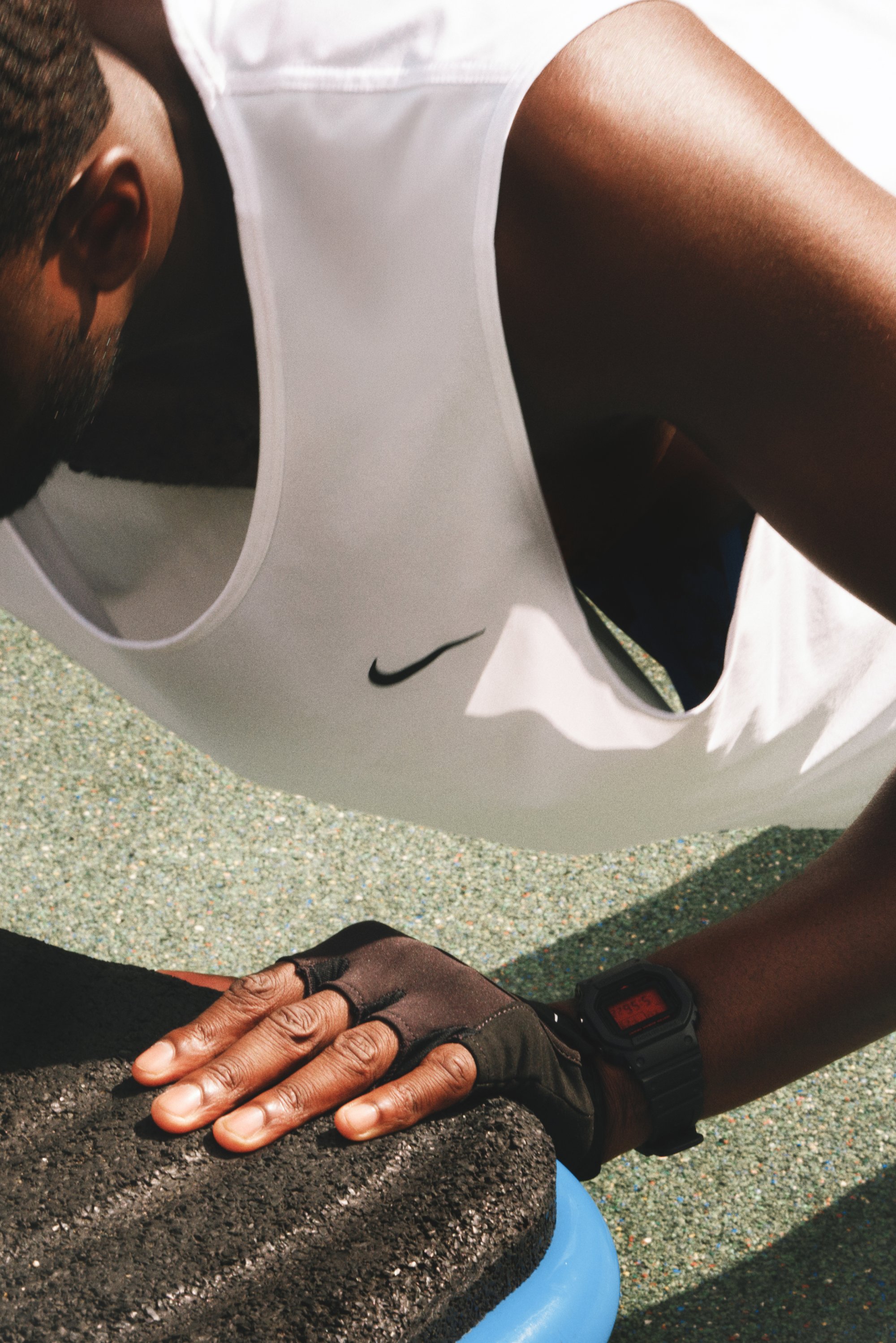 Zwarte fitnesshandschoenen die een textuur van een zwart oefenoppervlak vasthouden. Een digitale polshorloge toont de tijd. De persoon draagt een witte Nike tanktop.