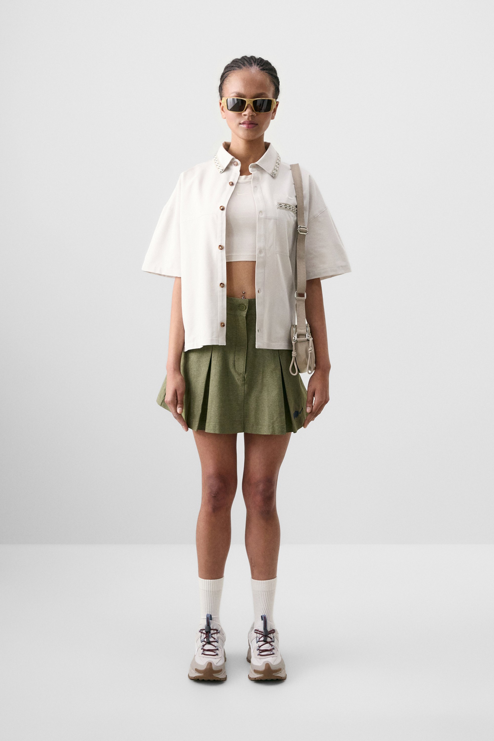 Una persona está de pie vistiendo una camisa blanca de manga corta, una falda plisada verde y zapatillas, accessorios incluidos gafas de sol y un bolso de hombro.