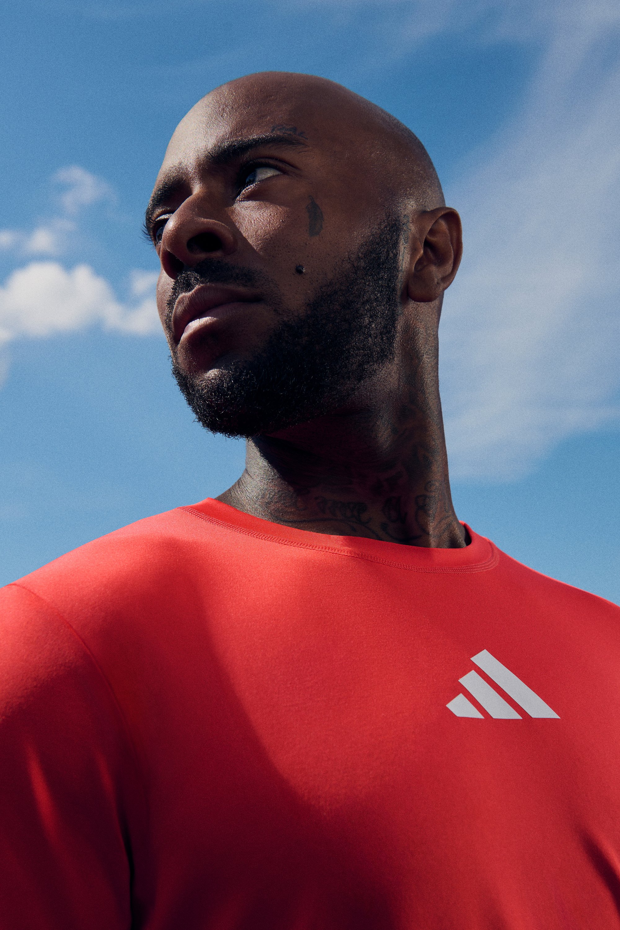 A man with a bald head and beard gazes upward, wearing a red shirt with the Adidas logo against a blue sky with clouds.