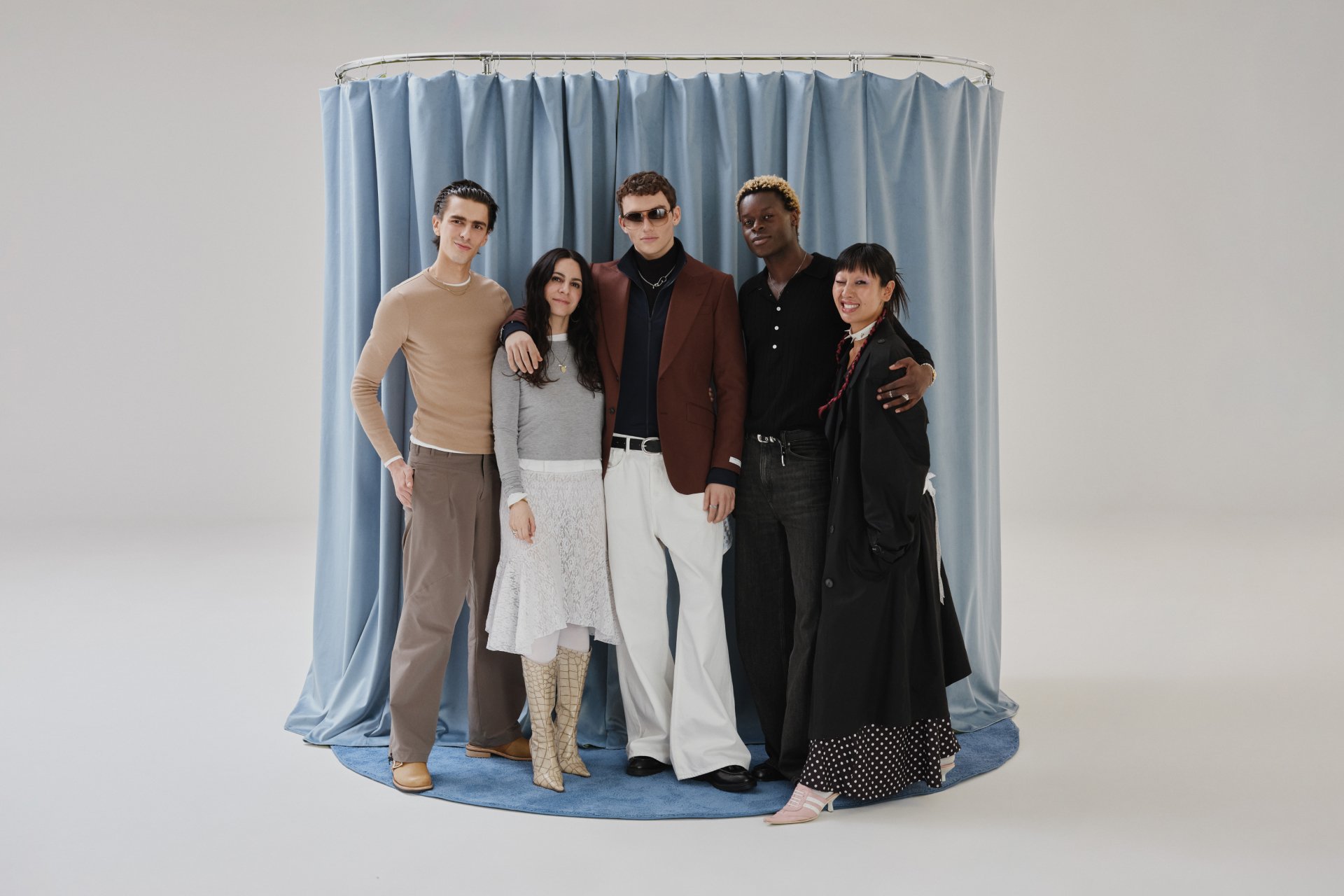 Five diverse individuals pose together in front of blue drapes. They sport stylish clothing and display friendly expressions.