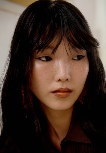 Femme aux longs cheveux foncés et aux taches de rousseur regarde sur le côté, portant un eyeliner foncé, un rouge à lèvres marron et des boucles d'oreilles rouges.