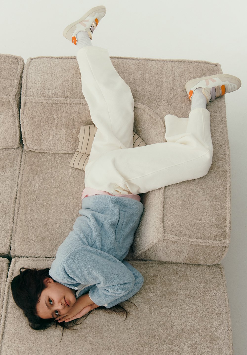 Chaqueta azul de forro polar con cremallera, pantalones de sudadera color crema y zapatillas con acentos en naranja, gris y rosa. Reposando sobre un sofá beige texturizado.