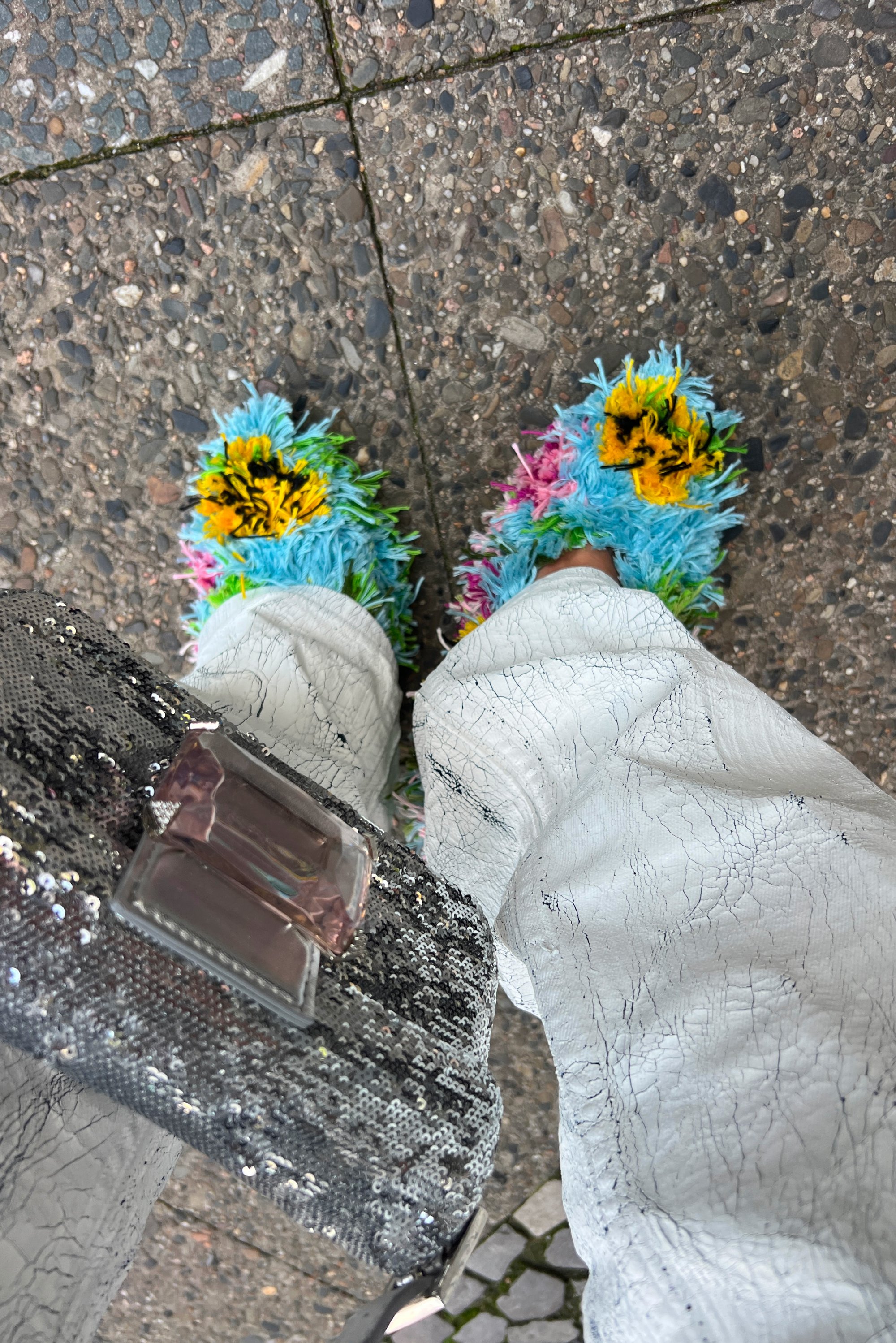 Piedi che indossano pantofole colorate e pelose con motivi floreali, pantaloni bianchi testurizzati e una borsa nera di paillettes su un pavimento di pietra.