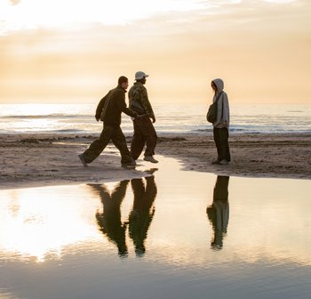 Twee personen wandelen langs het strand bij een reflecterend waterpoeltje tijdens de zonsondergang, terwijl een derde persoon in de buurt staat.