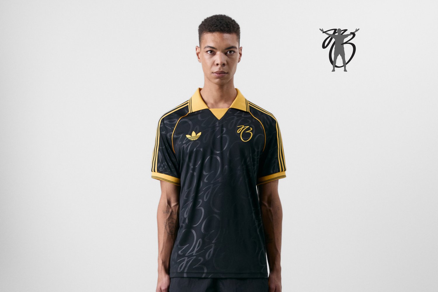Joven vestido con una camiseta deportiva negra con detalles amarillos y un logo, de pie frente a un fondo blanco liso.