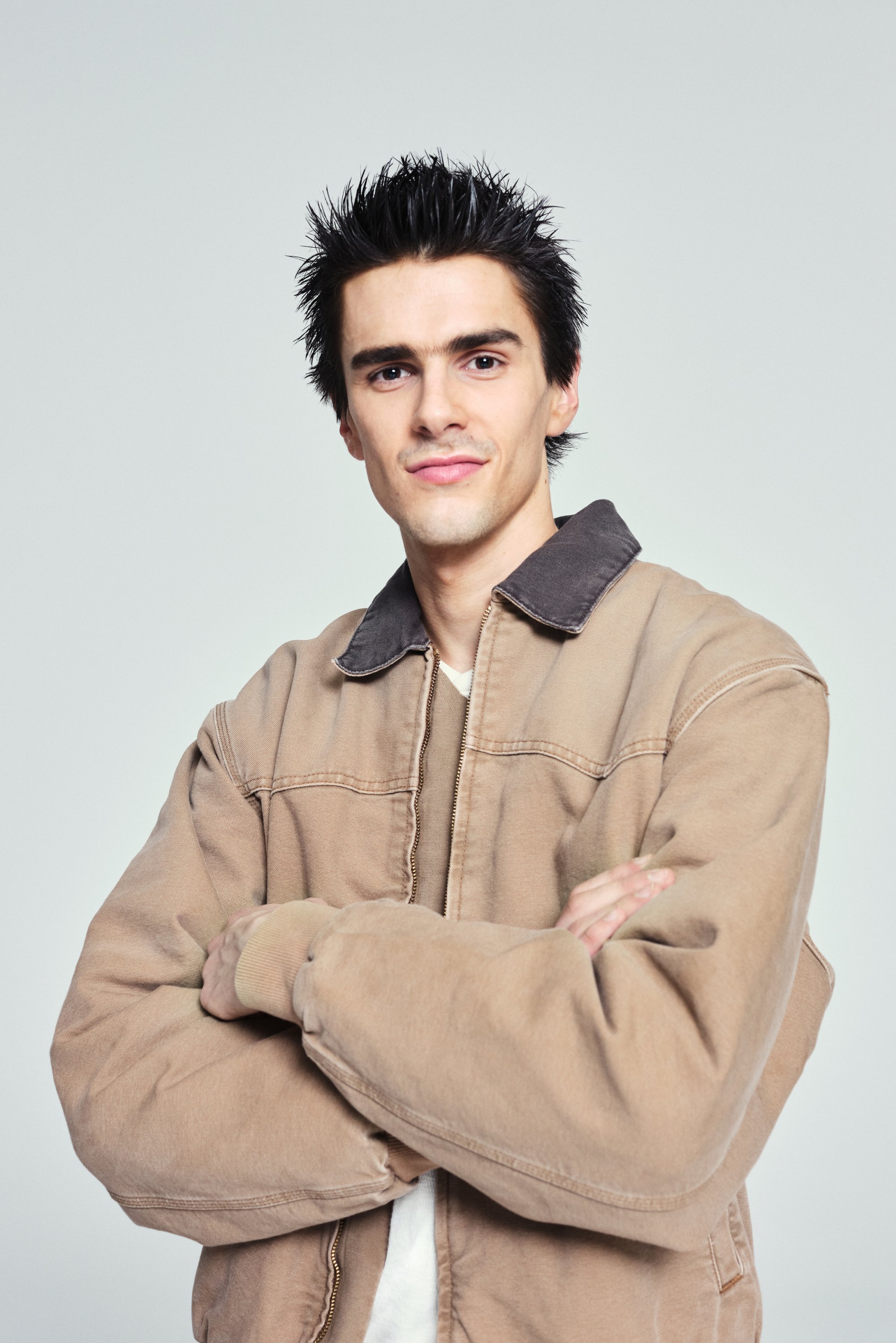 Young man with spiky hair stands confidently, arms crossed, wearing a beige jacket over a white shirt, against a light gray background.