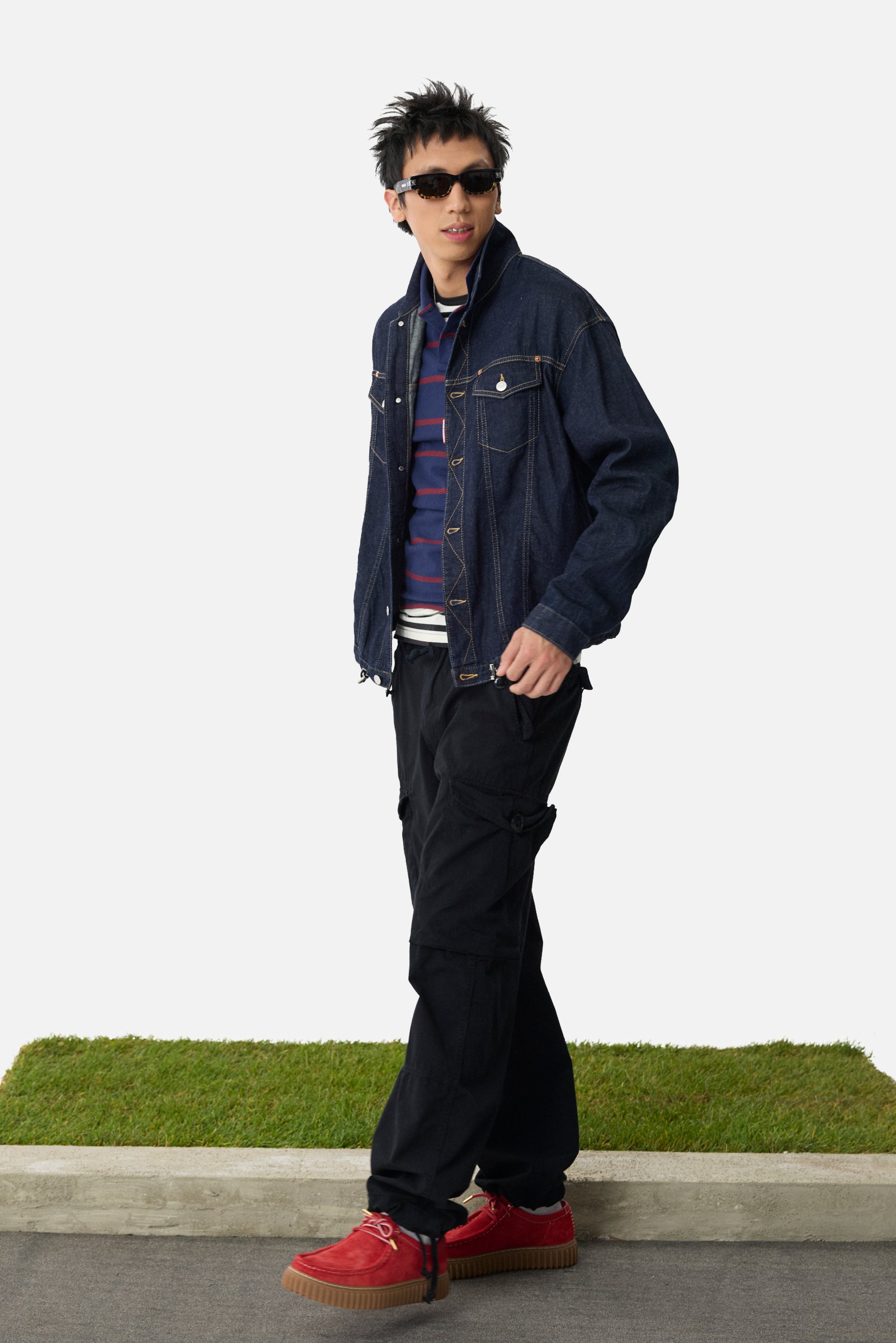 Chaqueta de mezclilla con dos bolsillos en el pecho, superpuesta a una camisa a rayas azul marino y roja, pantalones cargo negros y zapatos de plataforma rojos. Gafas de sol negras.