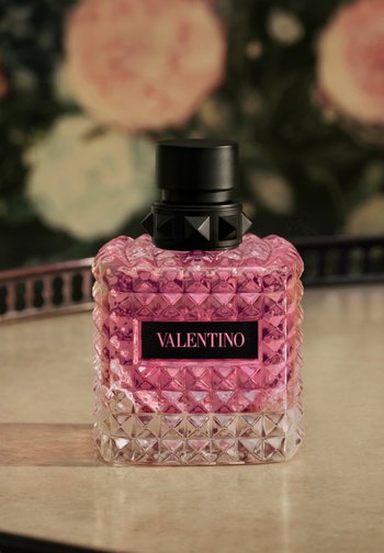 Kwadratowa różowa butelka perfum Valentino z czarną nakrętką z ćwiekami na lśniącej powierzchni i rozmytym kwiatowym tle.