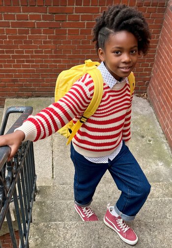 Enfant portant un pull rayé rouge et blanc, un sac à dos jaune et des baskets roses monte des escaliers extérieurs en tenant une rampe métallique noire.