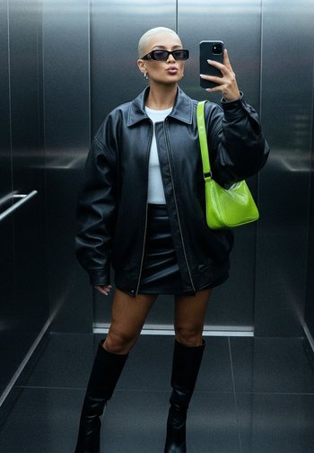Przesadzony czarny skórzany kurtka, biały top, czarna mini spódnica, wysokie czarne botki i błyszcząca zielona torebka. Lustrzane tło windy.