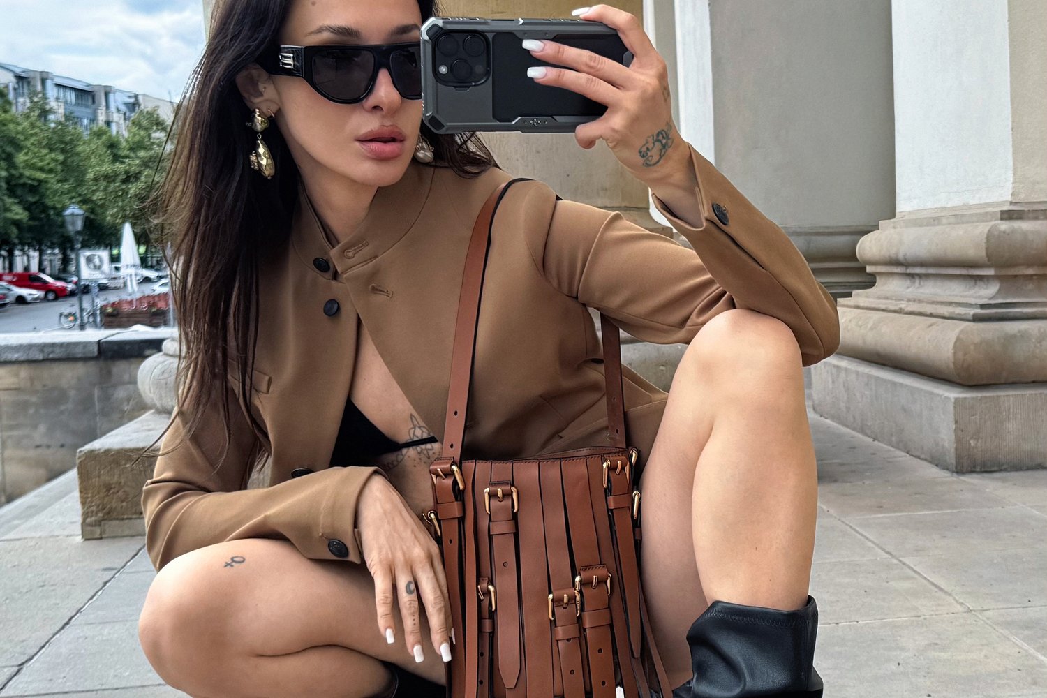 Blazer marrone su misura sopra un top nero, con una borsa a tracolla strutturata in pelle marrone dotata di manici e dettagli in oro, occhiali da sole neri e stivali fino al ginocchio.