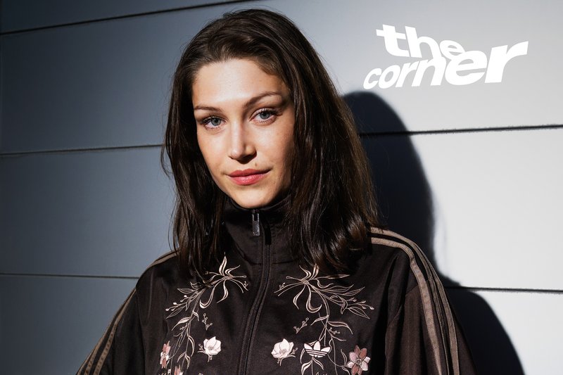 Young woman with dark hair wearing a black floral embroidered jacket stands against a light gray wall with shadow and text "the corner".