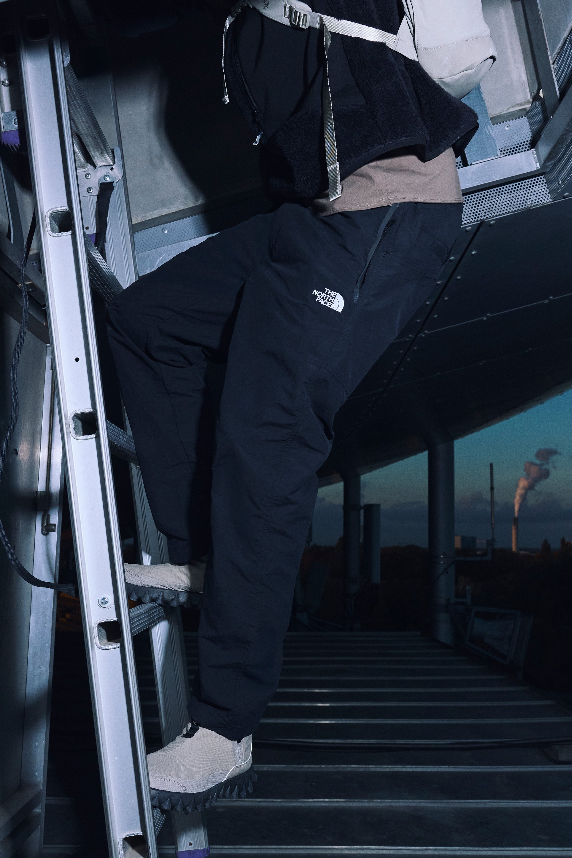 A person in dark pants and shoes climbs a metal ladder inside an industrial setting, with a smokestack visible in the background.