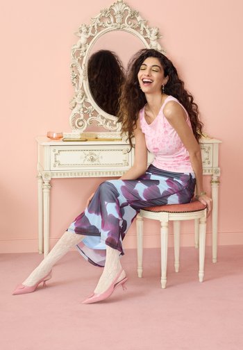 Femme aux longs cheveux bouclés souriante, assise sur un tabouret rembourré devant une coiffeuse vintage blanche avec un miroir orné, portant une jupe à motifs et des chaussures roses.