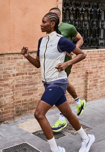 Δύο δρομείς, γυναίκα με λευκό φανελάκι και μπλε σκούρο κοντό παντελόνι, άνδρας με πράσινο πουκάμισο και μαύρο κοντό παντελόνι, τρέχουν σε ασφαλτοστρωμένο δρόμο της πόλης με φόντο τοιχο από τούβλα.