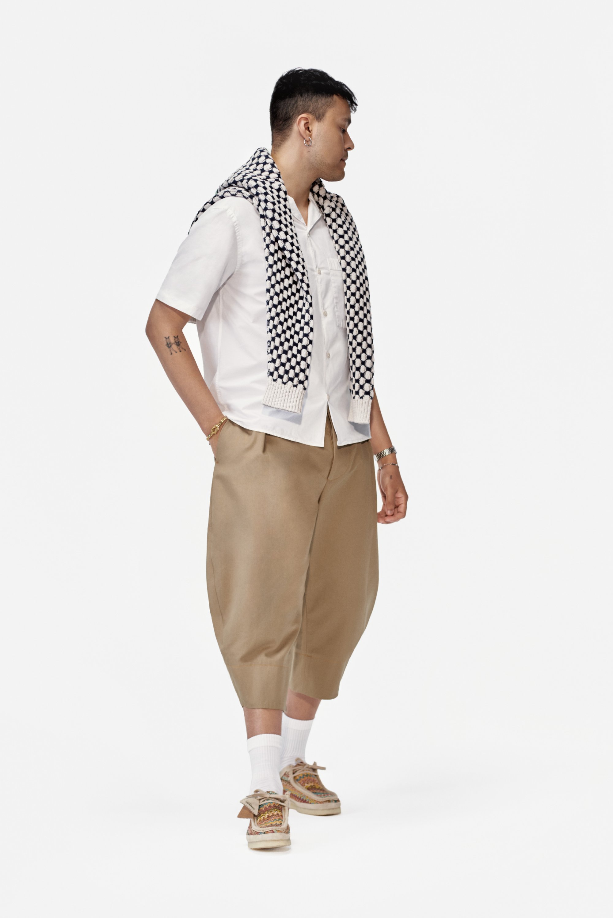 Una persona está de lado, vistiendo una camisa blanca de manga corta, pantalones beige de gran tamaño y zapatos con un patrón, con una bufanda estampada sobre los hombros.