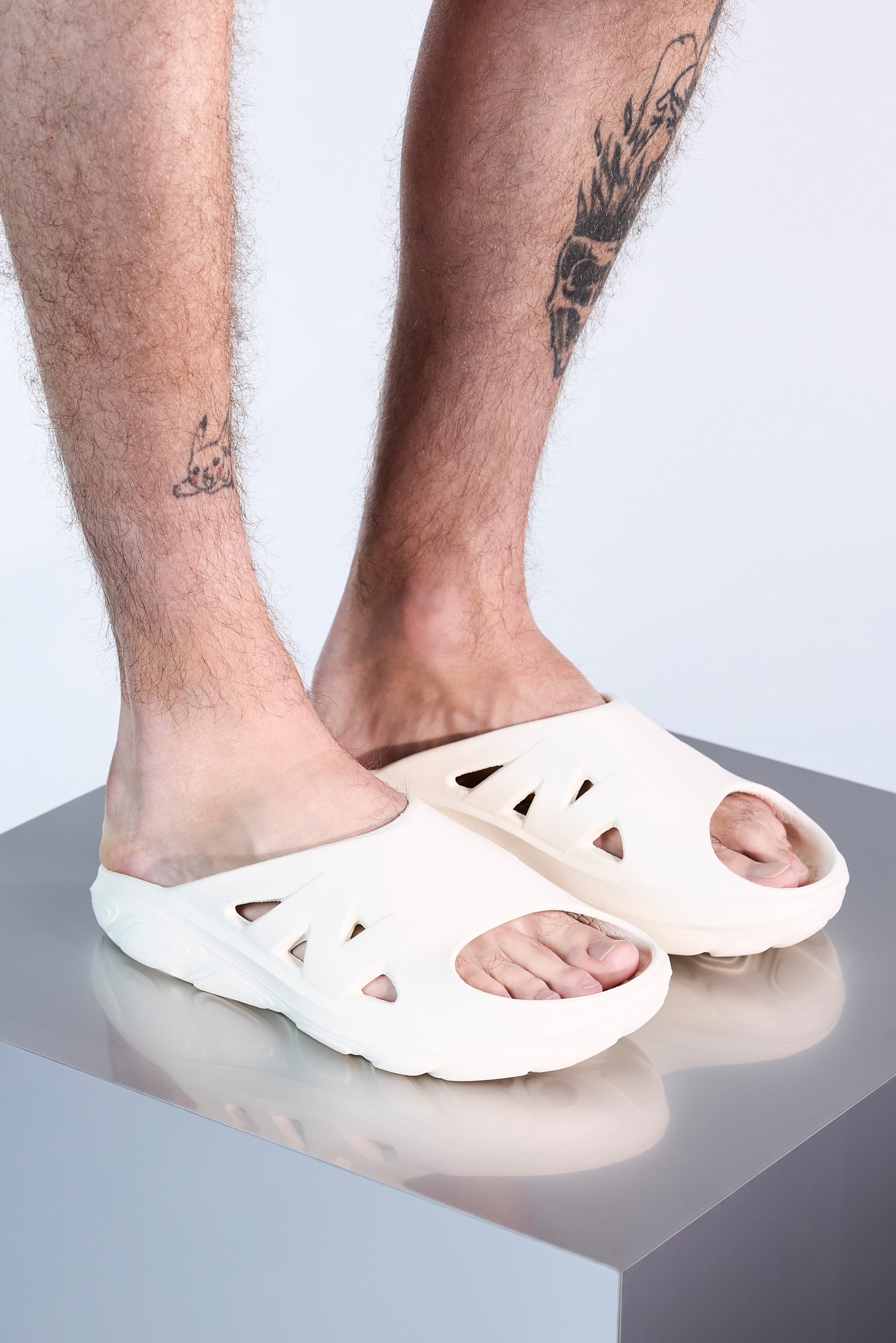 Feet in white slip-on shoes, showcasing tattoos on the shins. The shoes have cutouts and are placed on a smooth, reflective surface.
