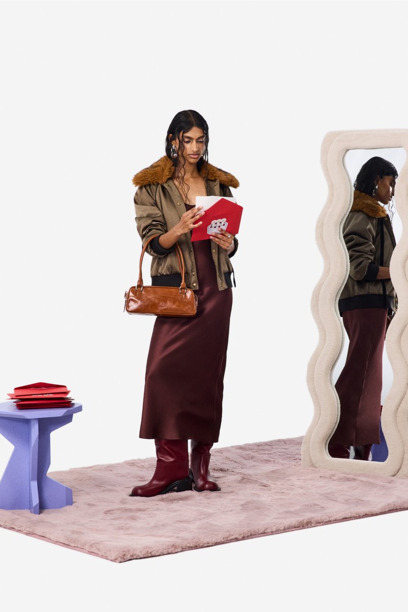Brun bomberjakke med imiteret pelskrave over en burgunder satin-natkjole. Tilbehør inkluderer en brun taske og en rød kuvert. Lilla sidebord og bølget spejl.