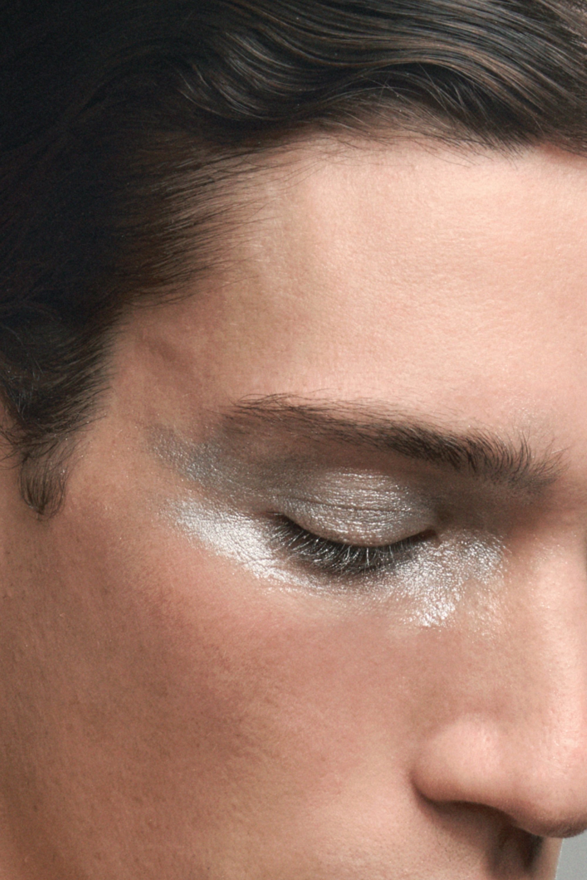 Close-up of a face featuring metallic silver eyeshadow, with a subtle expression, clear skin, and tousled hair.