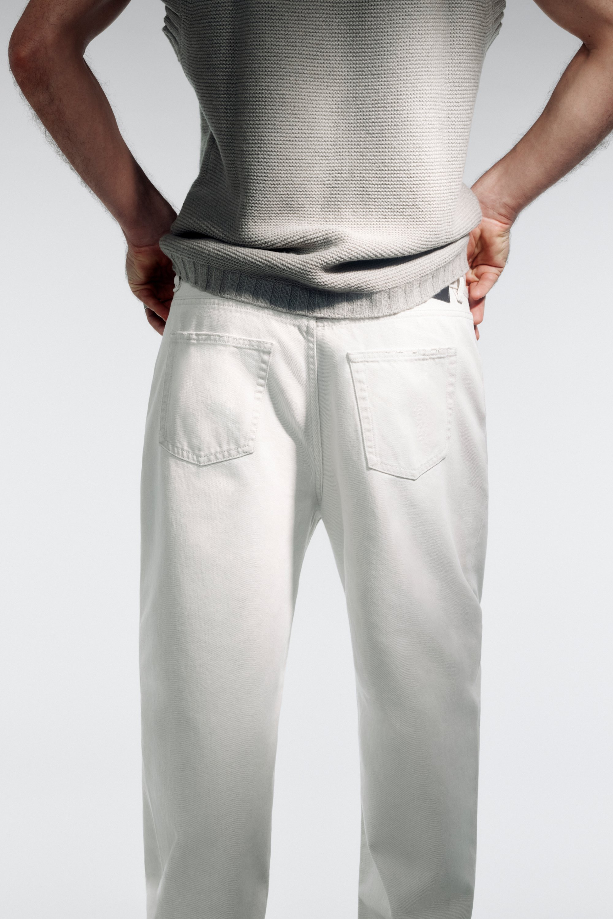 Una persona está de pie con las manos en las caderas, vistiendo un suéter gris y pantalones blancos, mostrando la parte trasera del conjunto contra un fondo claro.