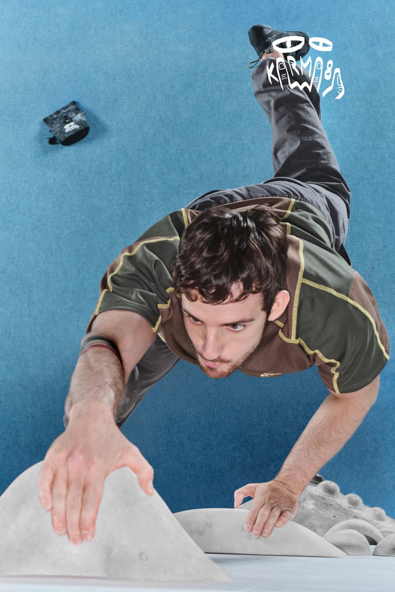 Hombre escalando una pared de boulder en interiores, extendiendo una mano hacia una presa de escalada, expresión de concentración, colchón de escalada azul debajo.