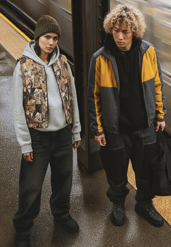 Avslappnade kläder: en i en brun camo-väst över en grå hoodie och vida jeans; den andra i en svart hoodie, grå och orange jacka och mörka byxor.