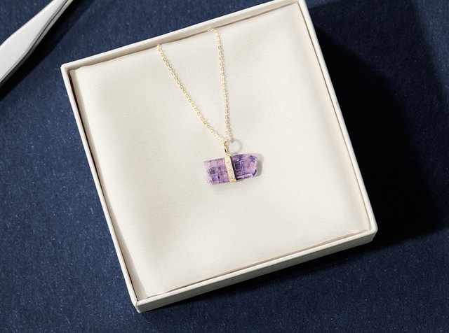 Gullkjede med en lilla krystallanheng, plassert på en myk beige pute inni en firkantet boks, med et enkelt kjededesign.